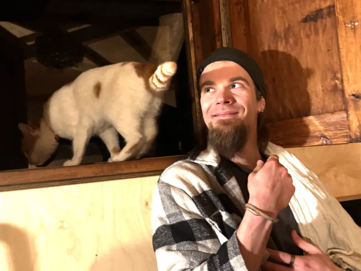 Dmitrii grinst zu Hause, während eine weiß-rote Katze über ihm durch eine offene Schrankklappe klettert