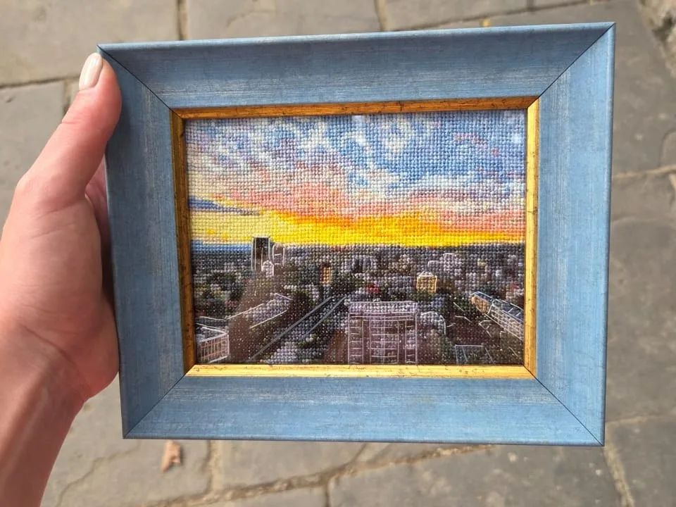 The finished cross stitch sunset in a blue and gold frame, held in one hand
